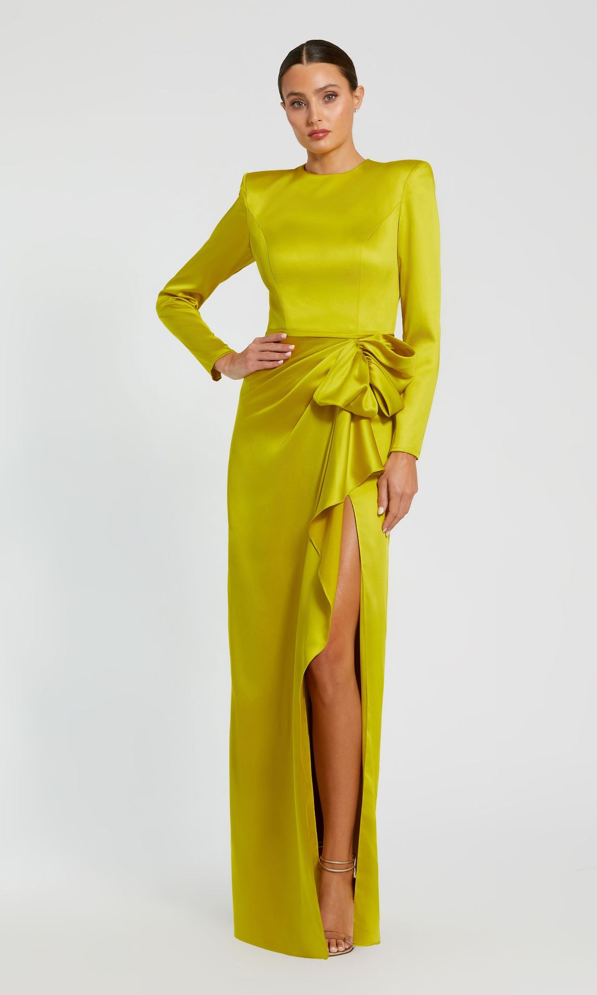 A woman models the Mac Duggal 26676 Bold-Shoulder Long Formal Dress—a bra-friendly, yellow-green gown featuring long sleeves, a side slit, and a draped waist bow—against a plain light background.